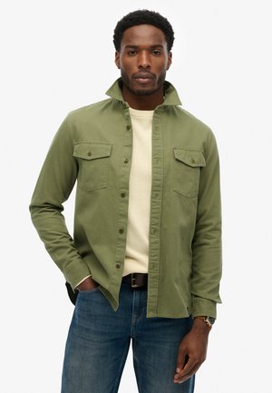 Olivgrünes Hemd zum Knöpfen mit zwei Brusttaschen, hochstehendem Kragen und langen Ärmeln. Trägt es über einem hellbeigen Hemd, kombiniert mit blauen Jeans.