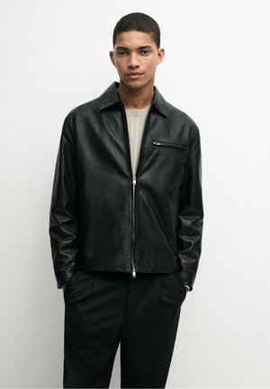 Jeune homme portant une veste en cuir noire zippée sur un pull beige clair et un pantalon noir, debout les mains dans les poches devant un fond uni.