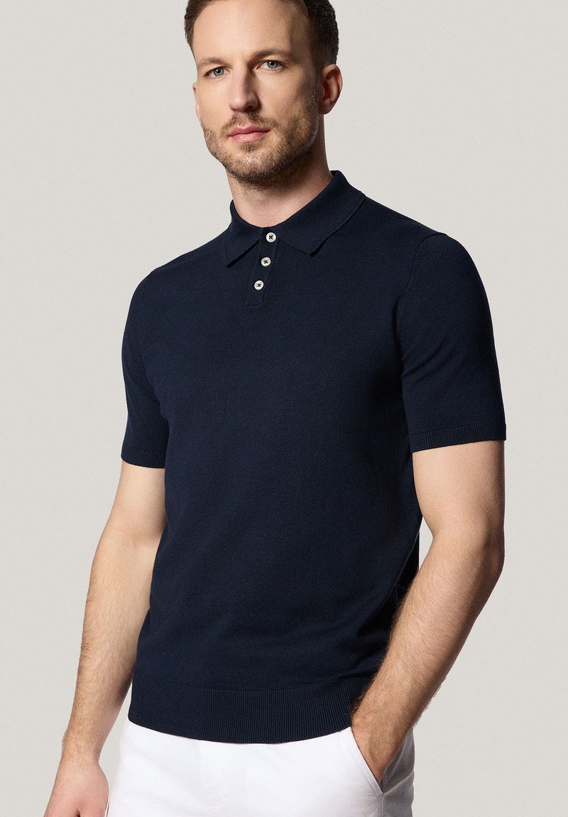 Granatowa koszulka polo z krótkim rękawem, wykonana z gładkiego materiału, z kołnierzykiem i zapięciem na trzy guziki, z dopasowanym talią i prążkowanymi mankietami.