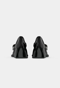 Paire de chaussures à talons en cuir verni noir brillant avec talons blocs et boucles latérales, vues de dos sur un fond blanc uni.
