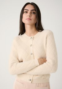 Cremfarbener Strickcardigan mit Rautenmuster, flauschiger Textur und silbernen Knöpfen. Verfügt über gerippte Bündchen und Saum, kombiniert mit gerippten Hosen.