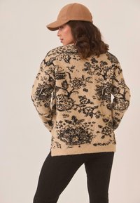 Suéter de punto beige con estampado floral y acentos negros, que presenta un corte holgado, mangas largas y una textura suave, combinado con jeans ajustados oscuros y una gorra.