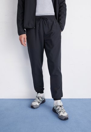 Personne portant un pantalon de jogging noir, des baskets blanches et grises, des chaussettes blanches et une veste noire par-dessus un t-shirt blanc côtelé, debout sur un tapis bleu.