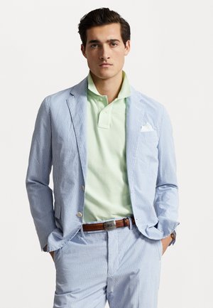 Costume pour homme en bleu clair avec des rayures blanches, associé à un polo vert menthe. Comprend un mouchoir de poche et une ceinture en cuir marron.