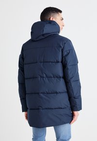 Zateplená tmavě modrá bunda s kapucí, s vodorovným prošíváním, dlouhými rukávy a mírně prodlouženým zadním lemem. Hladká textura.