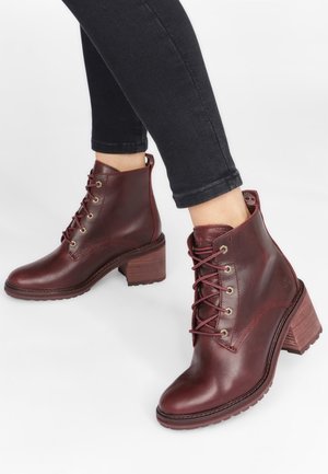 Personne portant des bottines en cuir rouge foncé à lacets avec talons carrés et un pantalon noir ajusté sur fond blanc.