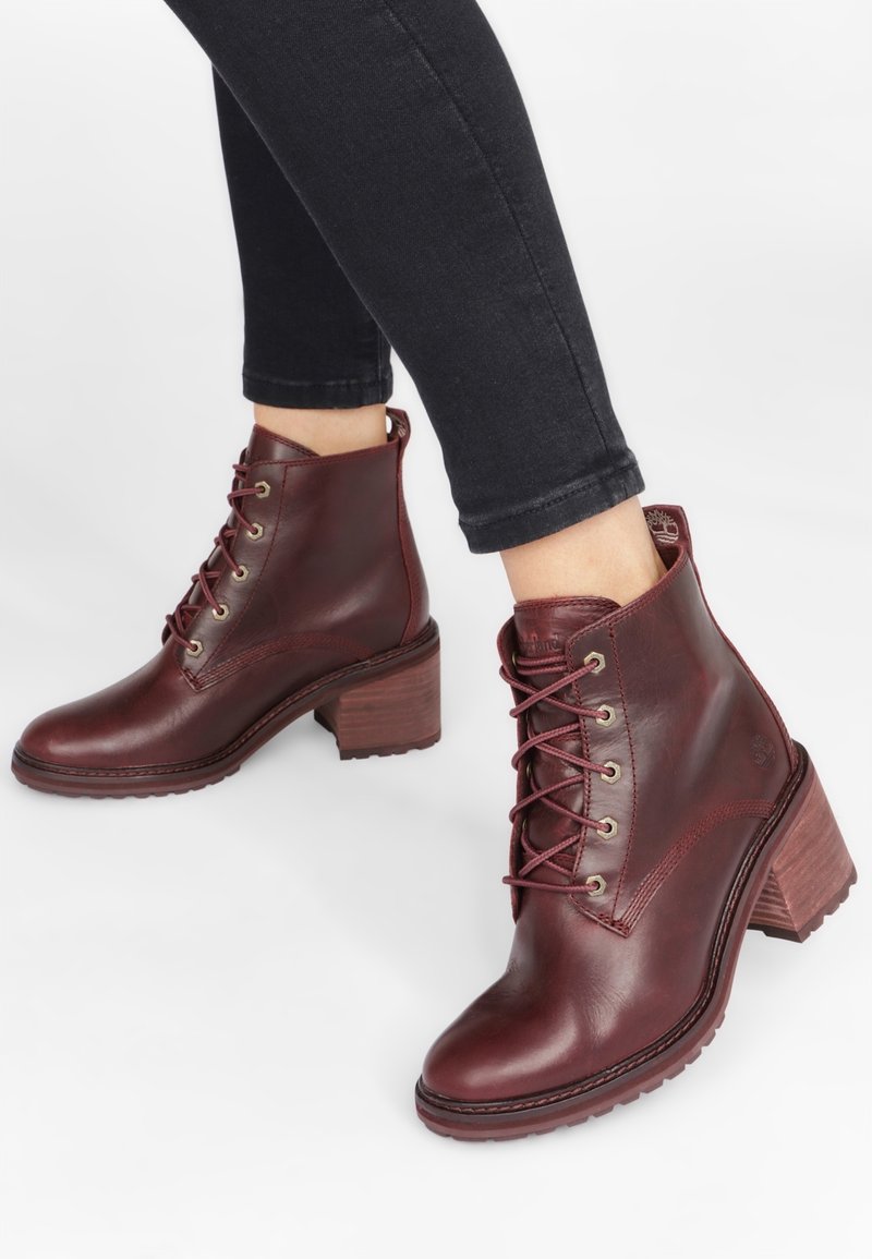 Personne portant des bottines en cuir rouge foncé à lacets avec talons carrés et un pantalon noir ajusté sur fond blanc.