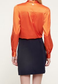 Blusa di seta arancione con maniche lunghe e polsini con bottoni, abbinata a una gonna aderente blu navy, caratterizzata da una leggera lucentezza e design sartoriale.