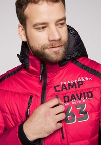 Chaqueta acolchada roja y negra con texto bordado, detalles de cremallera y un alto cuello. El material parece suave con una textura acolchada.