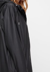 Veste noire imperméable avec une texture lisse et brillante, dotée d'une capuche, d'accents à cordon de serrage et d'une poche zippée sur le devant.