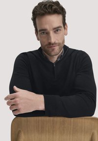 Schwarzer Pullover mit einem halben Reißverschlusskragen, aus weichem Strickstoff gefertigt. Mit langen Ärmeln und tailliertem Design, vor einem hellen Hintergrund präsentiert.