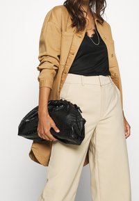 Bolso de cuero negro con grabado de cocodrilo sostenido en la mano con cierre de cordón. El modelo lleva una chaqueta marrón sobre una blusa negra y pantalones de pierna ancha color beige.
