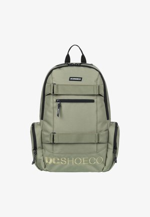 Mochila de tela verde con acentos negros, múltiples compartimentos con cremallera y correas ajustables. Presenta el logo "DCSHOECO" en dorado.
