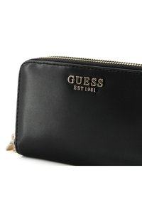 Portafoglio in pelle nera con logo del marchio in tono oro "GUESS EST 1981" e chiusura con zip, caratterizzato da una texture liscia e un design minimalista.