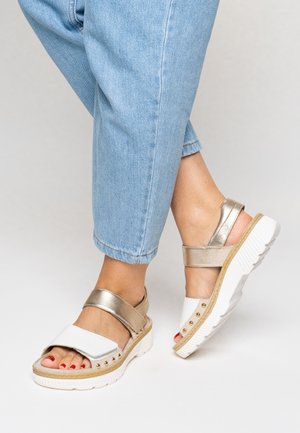 Weiße und goldene Sandalen mit zwei breiten Riemen, strukturierter Sohle und dekorativen goldenen Nieten auf der Plateausohle. Getragen mit hellblauen, gekürzten Jeans.