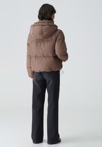 Brown padded jacket with a hood, featuring a soft fur lining around the collar. Worn with dark denim pants, viewed from the back.