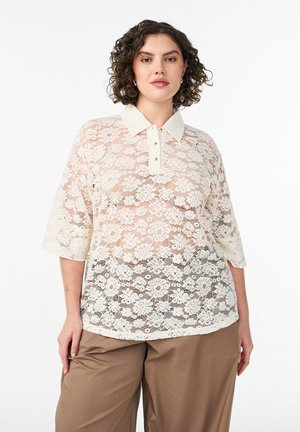 Žena s kučeravými vlasmi, oblečená v krémovej čipkovej kvetinovej krátkorukávovej košeli s golierom a hnedých nohaviciach s širokými nohavicami, stojaca pred bielym pozadím.