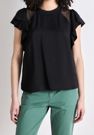 Kobieta w czarnej bluzce z falbankami na rękawach i siateczkowymi detalami, w połączeniu z zielonymi spodniami, stoi na tle jasnego, jednolitego tła.