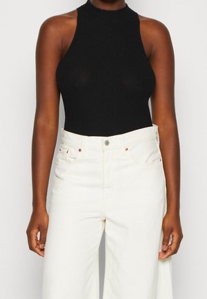 Personne portant un haut noir côtelé sans manches rentré dans un pantalon blanc taille haute à jambes larges, debout devant un fond uni.