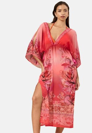 Nainen pukeutuneena vaaleanpunaiseen ja punaiseen kukkakuosiseen kaftaaniin, jossa on syvä v-kaula-aukko, leveät hihat ja korkea sivuhalkio oranssien bikineiden päällä.