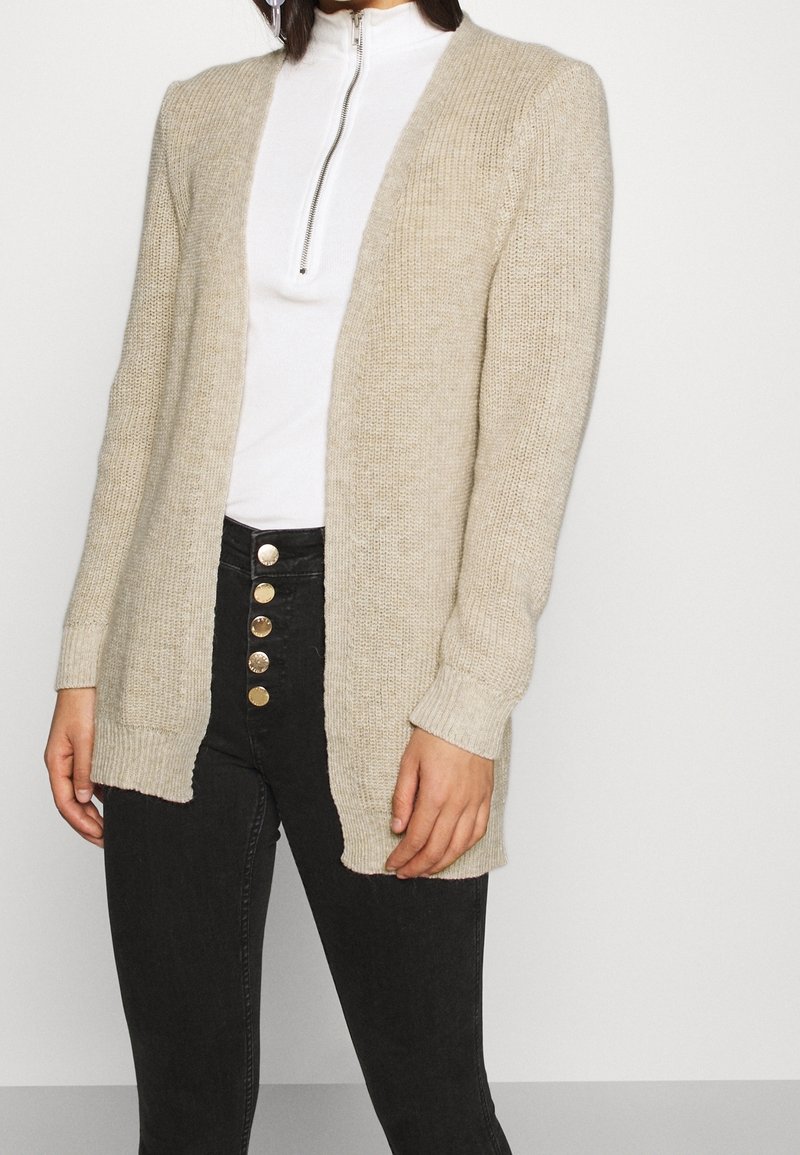 Jasnobeżowy sweter kardigan z dzianiny z otwartym przodem, żebrowanymi mankietami i bocznymi rozcięciami, zestawiony z białą bluzką z zamkiem przy szyi oraz czarnymi dżinsami z złotymi guzikami.