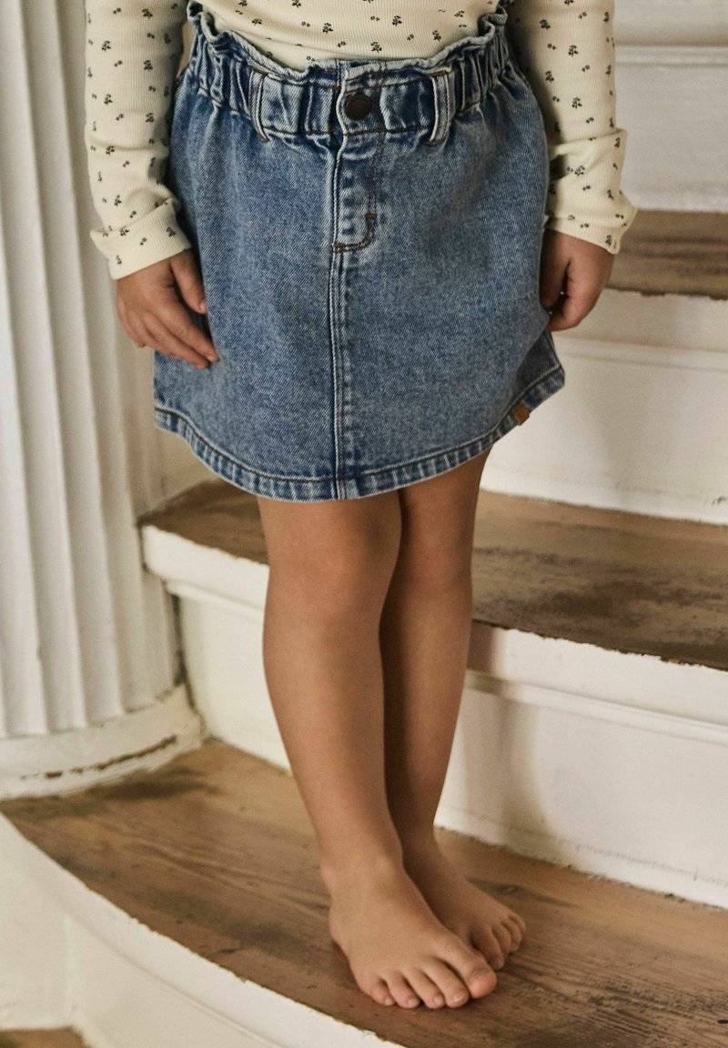 Enfant portant une jupe en jean bleue et un haut à manches longues crème avec un petit imprimé floral noir, debout pieds nus sur des escaliers en bois.