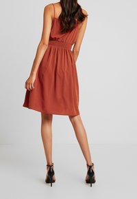 Šatové šaty v rzi, bez rukávů, s gumičkou v pase, splývavou sukní a hladkým povrchem. Nosené s černými sandály na vysokém podpatku.