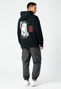 Hombre con sudadera negra con capucha con un gráfico de una mujer y el texto "OOPS I DON'T CARE", pantalones cargo grises y zapatillas blancas, de pie contra un fondo blanco.