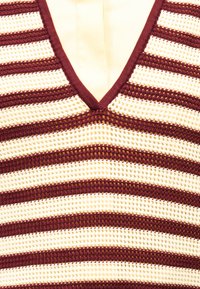 Κοντινό πλάνο αμάνικου πλεκτού τοπ με οριζόντιες ρίγες σε μπορντό και εκρού χρώμα και λαιμόκοψη σε σχήμα V.