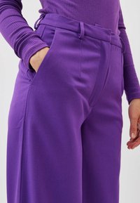 Pantalon large violet à taille haute, avec des plis à l'avant et des poches latérales. Le tissu est lisse avec une texture douce.