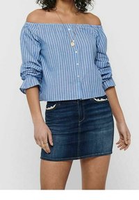 Femme portant un chemisier à rayures bleues et blanches, épaules dégagées, manches retroussées, et une mini-jupe en denim bleu foncé avec un bord en dentelle sur la poche.