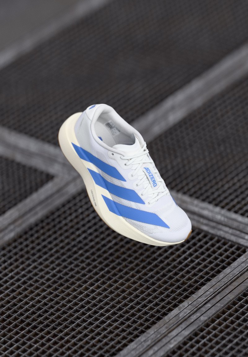 Hvid løbesko med blå striber og "ADIZERO" tekst, vist over en mørk metalrist.