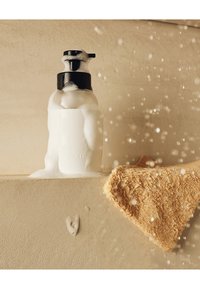 Witte zeepdispenser met zwarte pomp, overlopend met schuim, naast een gestructureerde beige handdoek, tegen een gladde beige achtergrond.