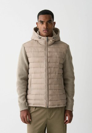 Beige vadderad jacka med huva, texturerade stickade ärmar, framdragkedja och horisontella sömmar. Lätt material, design för utomhusbruk.