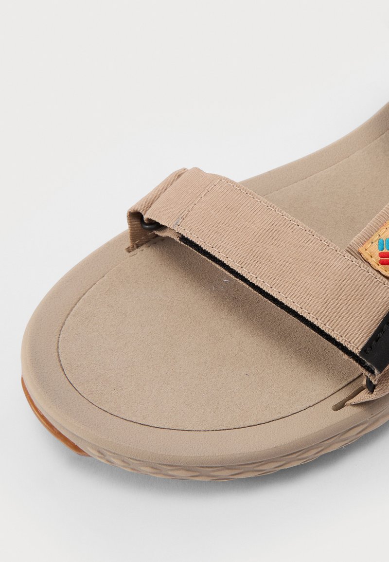 Beżowe sandały z miękką, teksturowaną wkładką, szerokim materiałowym paskiem oraz małym kolorowym logo. Płaska podeszwa z zaokrąglonymi krawędziami.