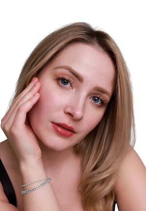 Primo piano di una persona con lunghi capelli biondo chiaro, pelle chiara e un sottile braccialetto d'argento. Texture della pelle liscia, trucco leggero con labbra di un rosso tenue.
