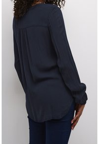 Frau mit lockigem Haar, die eine dunkelblaue Bluse mit langen Ärmeln und dunkelblaue Jeans trägt, steht vor einem einfarbigen hellen Hintergrund.