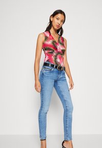 Fitted, sleeveless bodysuit with a vibrant pink and green palm leaf pattern, paired with light blue skinny jeans and a black belt.