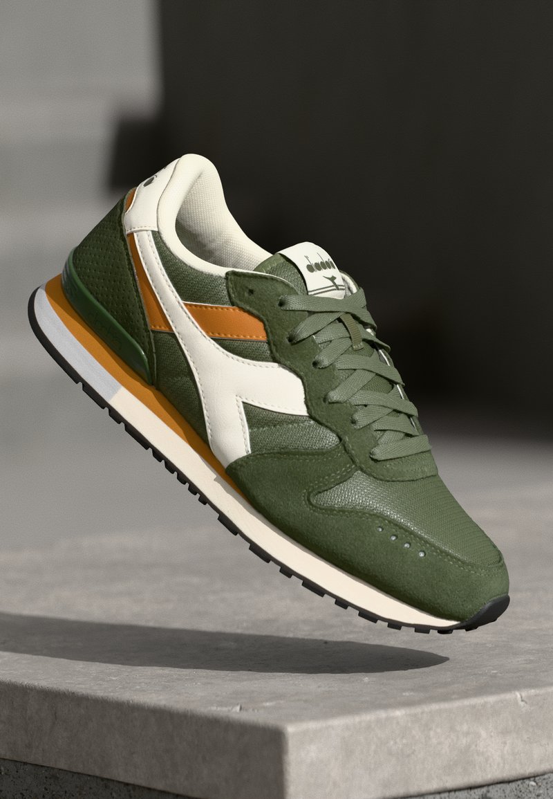 Green sneakers with white and orange accents, textured mesh and suede materials, featuring a lace-up design and a cushioned sole.