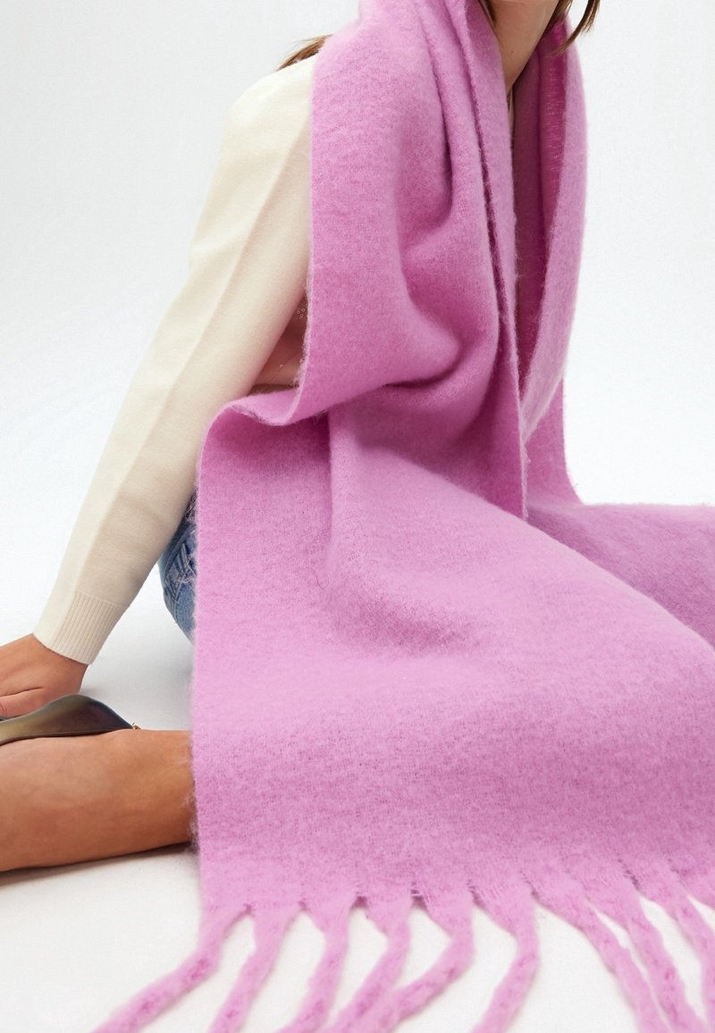 Personne assise portant un haut blanc à manches longues et un jean, enveloppée dans une grande écharpe ou un châle rose doux à franges.