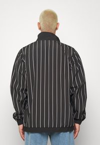 Svart jacka med vita vertikala ränder, med hög krage och elastiska ärmar, tillverkad av ett slätt, lätt material.