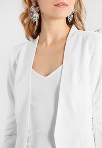 Žena v velkých stříbrných květinových naušnicích, bílém žebrovaném topu s V-výstřihem a bílém saku s nařasenými rukávy.