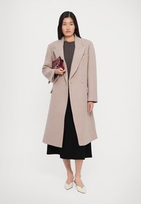 Woman wearing a beige long coat over a dark dress, holding a small maroon clutch, and wearing white pointed-toe heels, standing on a plain background.