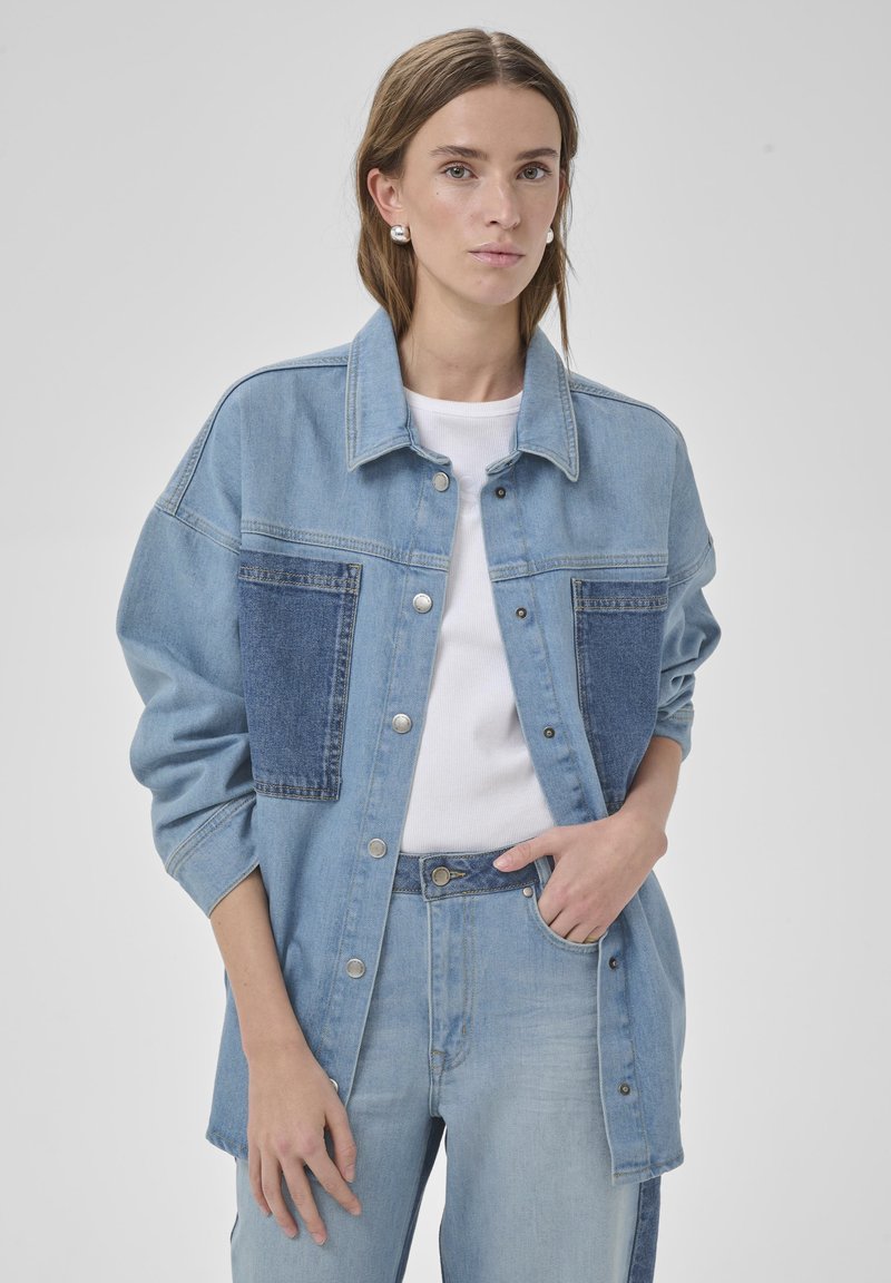 Junge Frau trägt eine hellblaue Jeansjacke mit dunkelblauen aufgesetzten Taschen und passende Jeans, steht vor einem schlichten Hintergrund.