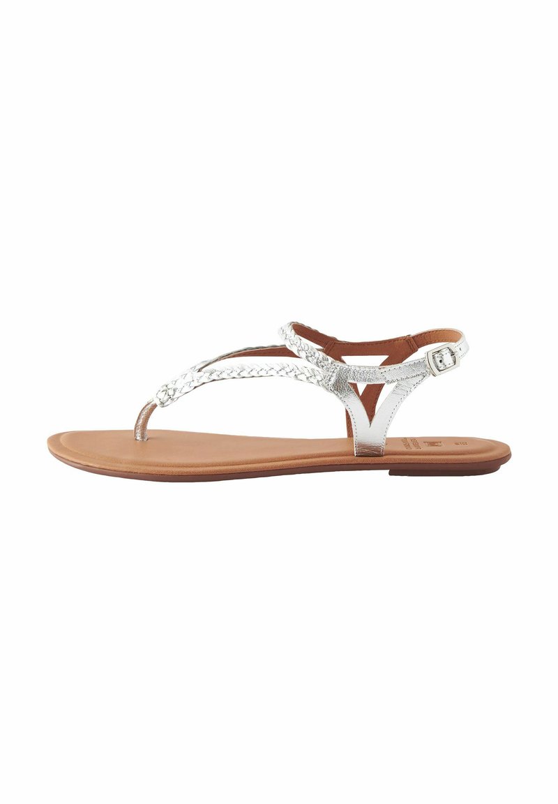 Sandali thong metallici argento con cinturini intrecciati, suola piatta marrone e fibbia regolabile alla caviglia per una vestibilità sicura.