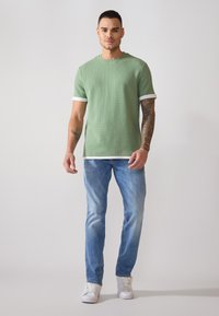 T-shirt verte à manches courtes texturée avec des bordures blanches, assortie à un jean bleu clair et des baskets blanches, présentée sur un modèle détenant une posture verticale.