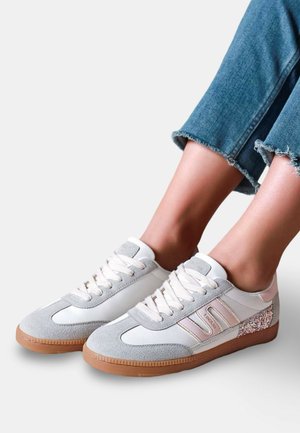 Personne portant des baskets gris clair et blanches avec des semelles en gomme et des détails floraux, assorties à un jean denim bleu raccourci.