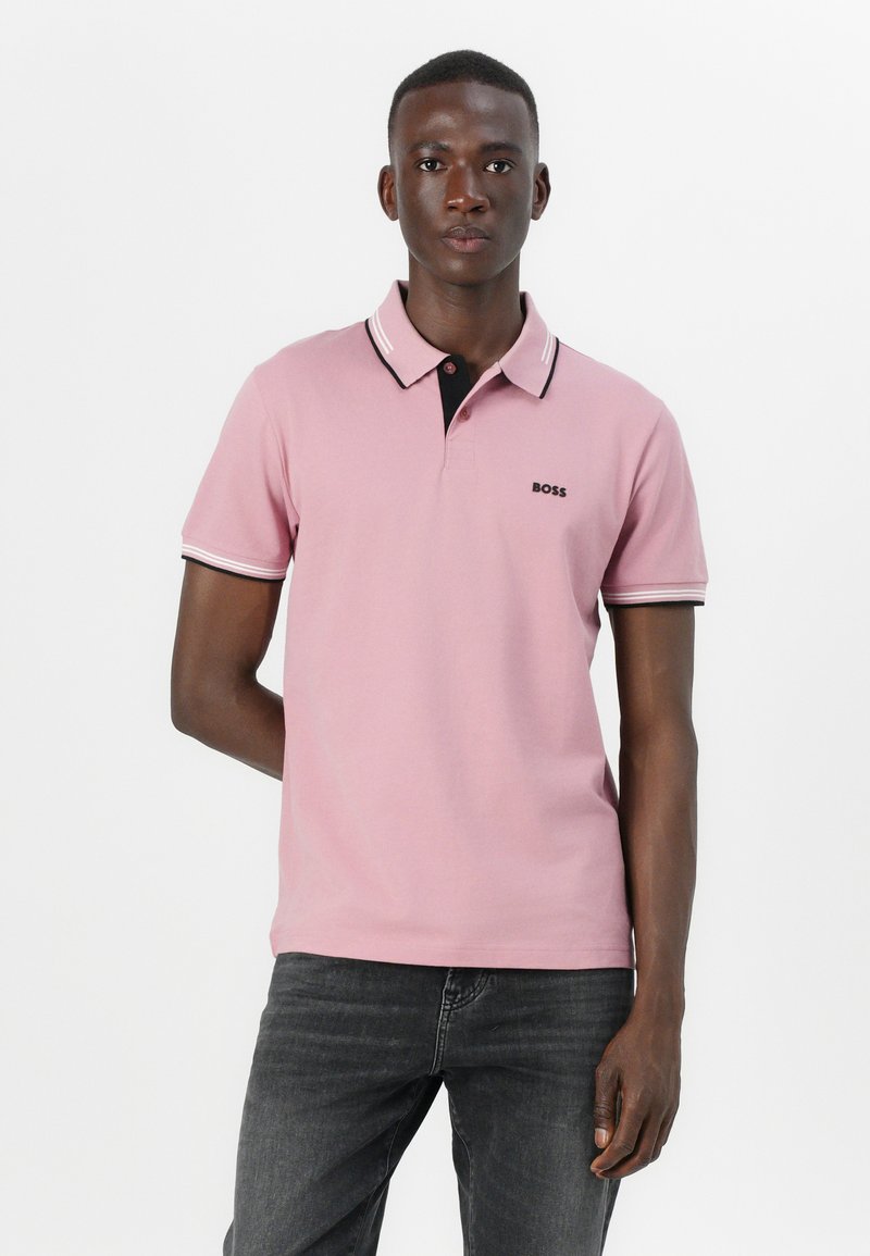Camisa polo rosa con un cuello negro y acentos blancos. Presenta un logo en el pecho y mangas cortas. Se lleva con jeans oscuros.