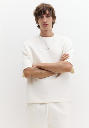 Fiatal személy rövid hullámos hajjal, teljesen fehér alkalmi ruházatban, ezüst nyaklánccal és kar tetoválásokkal, karját keresztbe téve, egyszínű háttér előtt állva.