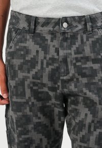 Nærbilde av en person som har på seg grå og svart pikselert camouflage-bukser med knappefal, hvor hånden er delvis synlig.